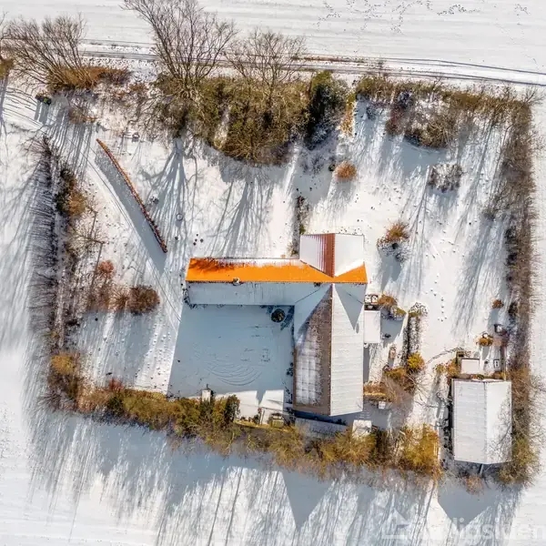 Hus med rødt tag ses ovenfra omgivet af træer og sneklædte marker. Skovområde omkranser husets område, mens skygger fra træerne strækker sig over jorden i vintersolen.