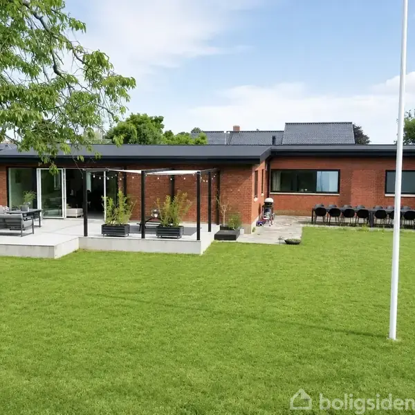 Rød murstenshus med store vinduer står stille i en velplejet have med grøn græsplæne og moderne terrasse med havemøbler. En sving hænger fra et træ til venstre.