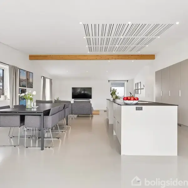 Et moderne køkken-alrum har et hvidt køkkenbord med røde æbler, omgivet af grå stole og bord. En sofa og fladskærms-tv er placeret i baggrunden.