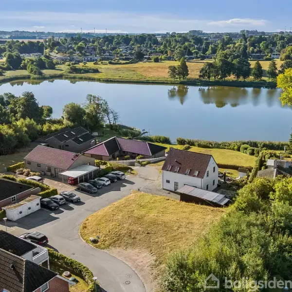 Huse ligger stille ved en sø omgivet af frodige træer og græs. Flere biler er parkeret ved siderne af husene, mens søen strækker sig i baggrunden under en blå himmel.