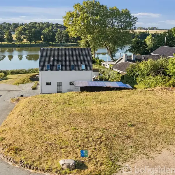 Hus beliggende ved en sø med spejlblankt vand. Foran huset er en græsplæne, og til venstre ses parkerede biler. Baggrunden består af træer og landskab.