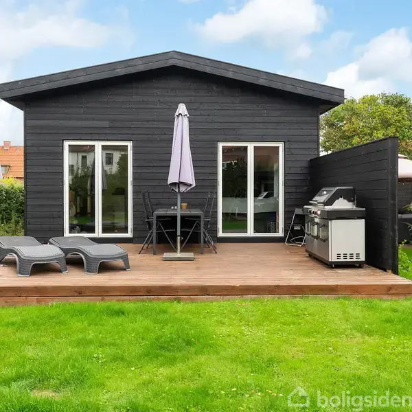 Sort træhus med to glasdøre åbner ud mod en træterrasse. Terrassen har liggestole, et rundt bord med parasol, og en grill. Græsplæne og grønne buske omringer huset.