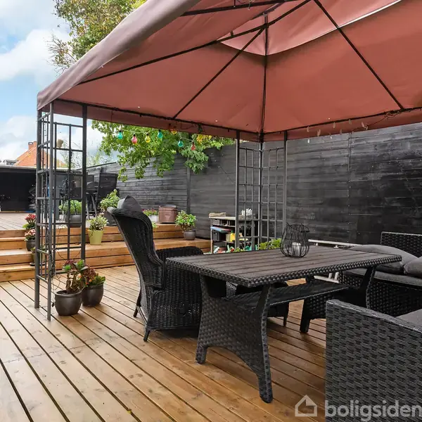 En terrasse med et brunt havepavillon, indeholdende sorte flettemøbler. Omkring er der blomster og potteplanter, og en trappe fører op til et andet niveau med en cykel og skure.