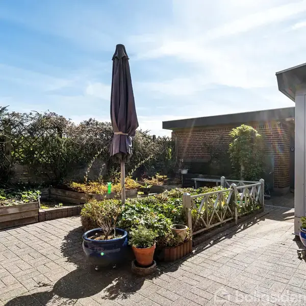 Terrasse med blomsterkrukker og en stor parasol. Planter vokser i højbede, og en lav mur omgrænser området. En glasdør fører ind til huset under en klar blå himmel.