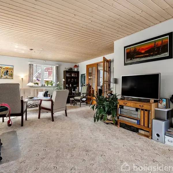 En stue med tæppebelagt gulv, indeholdende en sofa, bord, og stole. Et fjernsyn står på et skab foran en væg med billeder. Store vinduer giver naturligt lys til rummet.