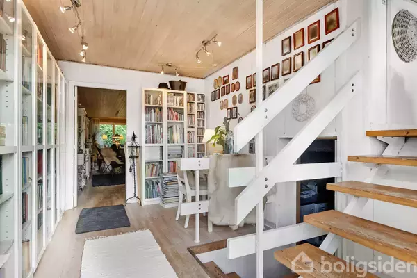 Trapper med trætrin fører opad ved siden af et lyst rum med mange bogreoler og et hvidt skrivebord. Væggene er dekoreret med indrammede billeder.