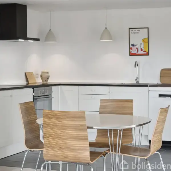 Rundt spisebord med fire træstole står i et moderne, hvidt køkken med sorte bordplader. To vedhængslamper hænger over bordet. Væggen har indrammede grafiske prints.