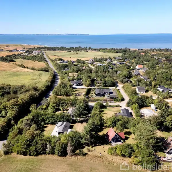 Landsby omgivet af grønne marker og skove, strækker sig mod en blændende blå kystlinje i det fjerne. Veje snor sig gennem området med spredte huse beliggende i landskabet.