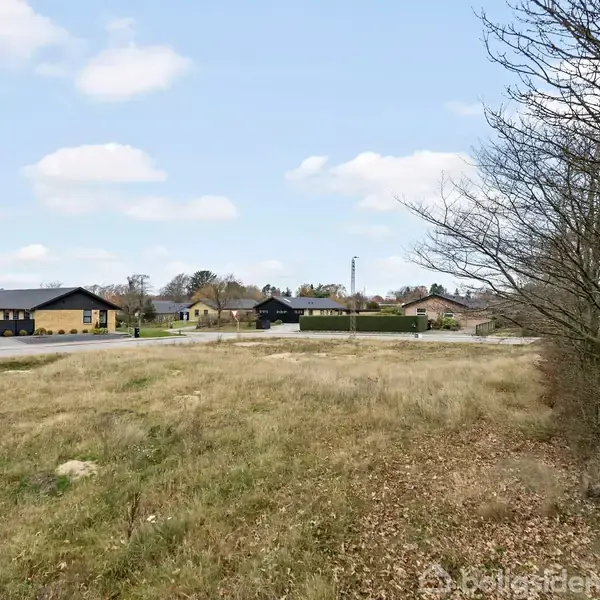 Græsareal breder sig til venstre, omkranset af en lav hæk til højre. Boligområde med lavthængende huse i baggrunden under en klar himmel.