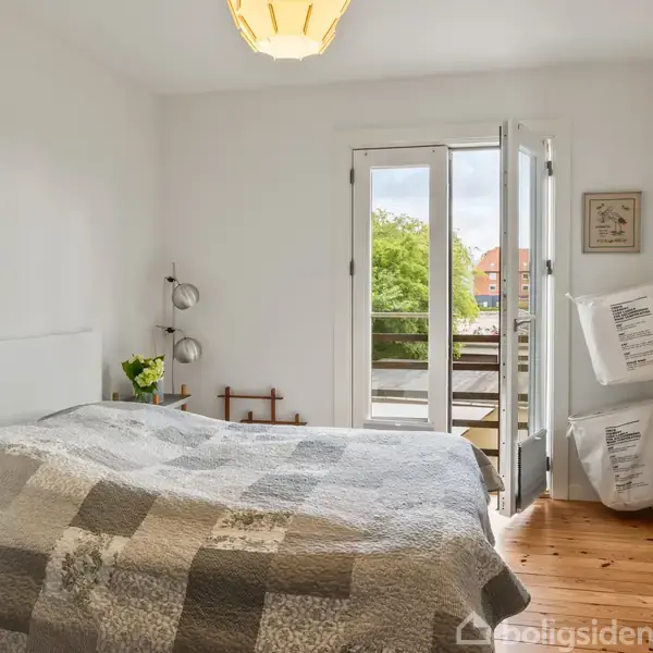 Soveværelse med seng dækket af patchwork-tæppe, to puder ved siden af, og døre, der åbner til en balkon med grønne træer udenfor. En lampe hænger fra loftet, og der står en bog ved siden af sengen.