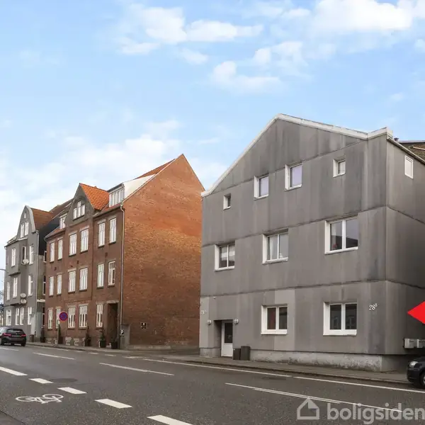 Boligbygning i tre etager med gråt facade, står langs en byvej. Røde murstenshuse ses ved siden af. En parkeret bil og cykelsti er synlige i forgrunden. Rød pil peger mod bygningen.