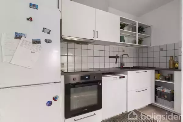 Et funktionelt køkken med en ovn, opvaskemaskine og køleskab. Køleskabet har magneter og papirer. En vask er placeret under åbne hylder med planter. Flisevægge skaber et rent look.