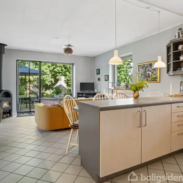 Køkken med lys træbordplade, hvor en vase står. I baggrunden er spisebord med stole, en brændeovn og en dør til terrasse. Lysindfald fra vinduer og billeder på væggen.