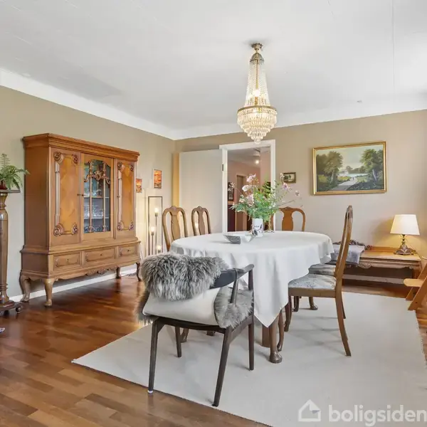 Spisebord dækket med dug står centralt i en elegant spisestue. Der er omgivet af klassiske træmøbler, kunstmaleri på væggen, lysekrone i loftet og trægulv.