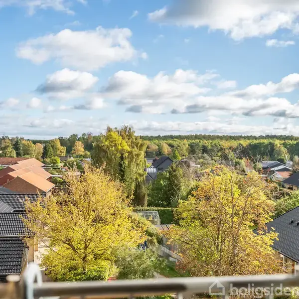 Et åbent vindue viser huse og træer med efterårsfarver, set fra en højde. Skyer spredt over en blå himmel danner baggrund, hvilket skaber en fredfyldt forstadsudsigt.