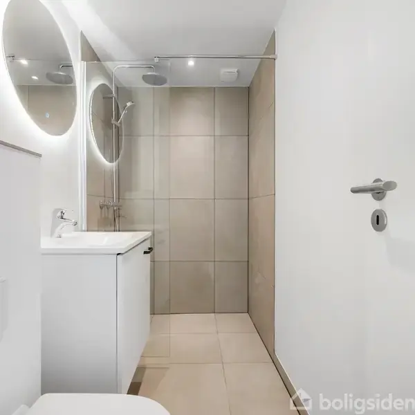 Et moderne badeværelse har en bruser med glasvæg, et rundt spejl med baggrundsbelysning over en vask, og et toilet. Der er lyse vægge og fliser, der skaber et minimalistisk udtryk.