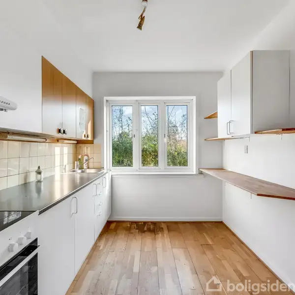 Et lyst køkken med hvide skabe, træhylder og en sort kogeplade. Rummet har trægulve og et vindue i baggrunden, der lukker naturligt lys ind og viser grønne træer udenfor.