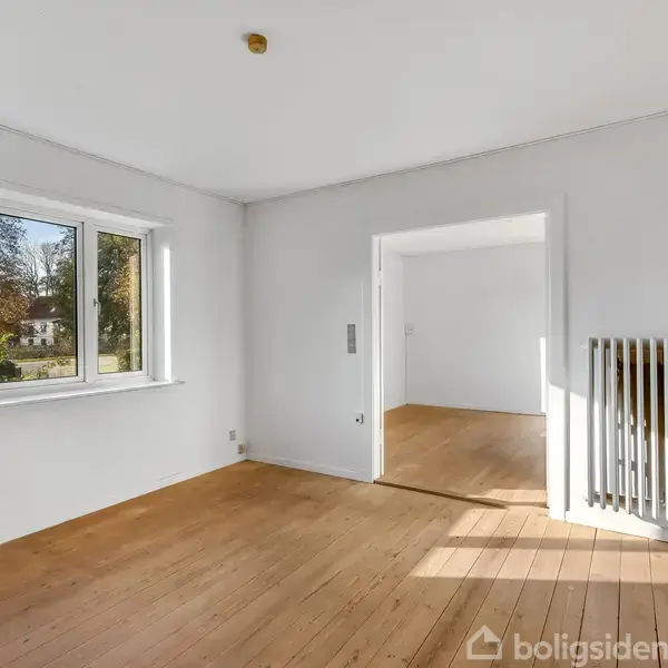 Et tomt rum med trægulv, hvor en radiator er monteret på væggen. Et stort vindue viser en have udenfor. Døråbning fører til et andet rum.