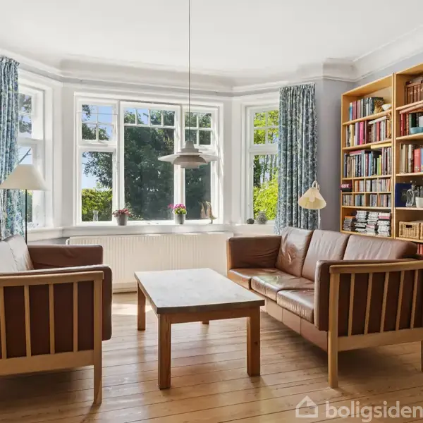 To sofaer og et sofabord står i en lys stue med store vinduer og masser af naturligt lys, omgivet af bogfyldte reoler og gulvlamper.