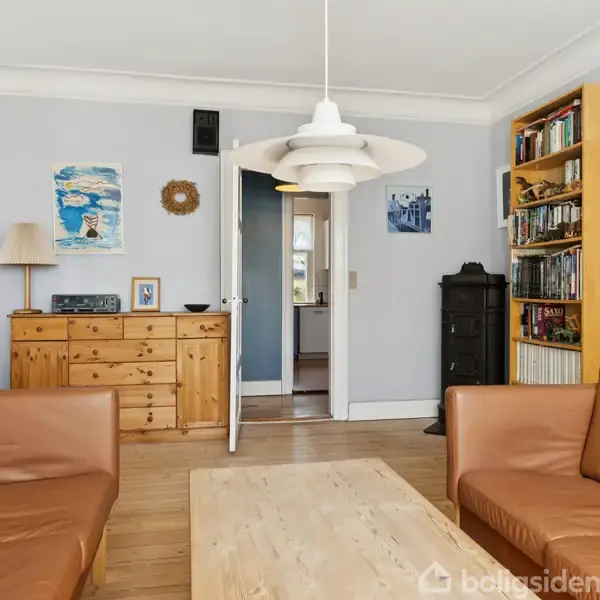 Brune lædersofaer står rundt om et træbord i en stue med lyseblå vægge. En kommode og bogreol står ved væggene, mens en lampe hænger over bordet. Malerier hænger på væggen.