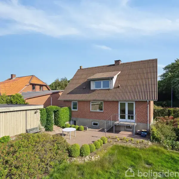 En murstensvilla med tagvindue står stille i en grøn have med terrasse. Baghaven omkranses af buske, og nabohuse er synlige under en blå himmel.
