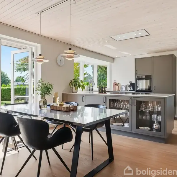 Spisebord med sorte stole står i et lyst køkken med træloft og vinduer mod en terrasse og have. Midt på bordet er en plante, og der hænger lamper over bordet.