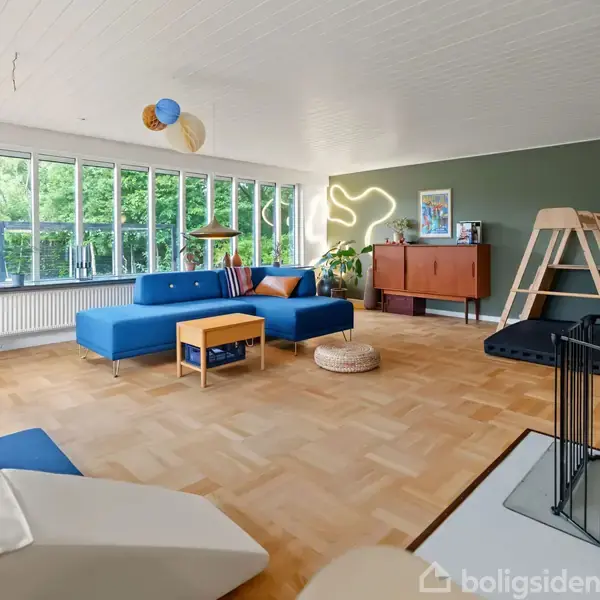 En stor, lys stue med moderne møbler udforsker hygge. En blå hjørnesofa står foran store vinduer med udsigt til grønt. Et dekorativt lysdesign hænger på en grøn væg.