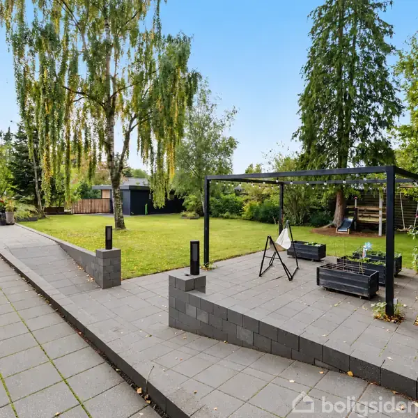 En have med en flisebelagt terrasse, hvor der står en sort pergola og grå sofaer. Græsplæne og træer omkranser området, og der er et lille skur i baggrunden.