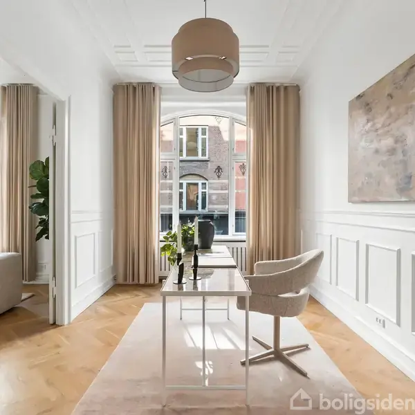 Et moderne værelse med et bord og stol står foran store vinduer med gardiner. Væggene er hvide med paneler, og et maleri hænger til højre. En plante er i hjørnet.