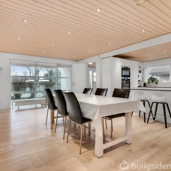 Spisebord med sorte stole står i et lyst rum med trægulv og hvidt loft. Ved siden af bordet er der et moderne køkken og store vinduer mod en have.