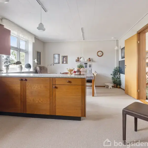 Køkkenområde med træskabe, stille og tomt. Bordet er i baggrunden, dekoreret med genstande. Rummet er lyst med store vinduer og neutral indretning. Vinduet giver udsigt til grønne omgivelser.