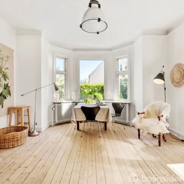 Et lyst rum med trægulve har et bord og stole foran en karnap med store vinduer. Rummet er dekoreret med en vægplakat, en gulvlampe og en flettet kurv.