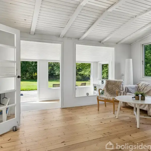 Et sommerhus med blå tag beliggende i en græsplæne omgivet af træer. To små sorte udhuse står ved siden af. En træbænk og borde ses på terrassen foran huset.