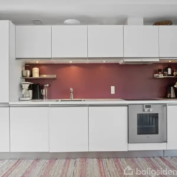 Sort lædermøblement står i en stue med åbent køkken og spiseplads. Klokken på væggen viser tidspunkt. Store vinduer giver udsigt til grønne omgivelser. En gang leder til et soveværelse.