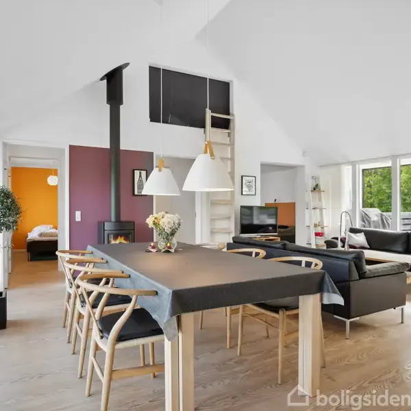 Spisebord med stole står i stue med brændeovn og lamper, flankeret af sofaer. Rummet har store vinduer med udsigt til grøn natur og en dør mod et værelse.