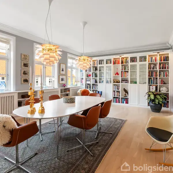 Et spisebord med stole er omgivet af en væg af bogreoler fyldt med bøger. To moderne lamper hænger over bordet. Rummet har lyse trægulve og store vinduer.