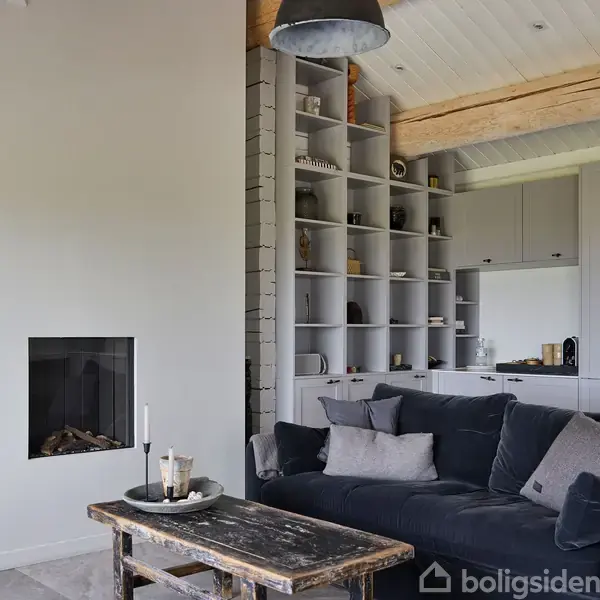Reol med dekorative genstande står ved siden af en pejs i en moderne stue, hvor en mørk sofa med puder skaber en hyggelig atmosfære.