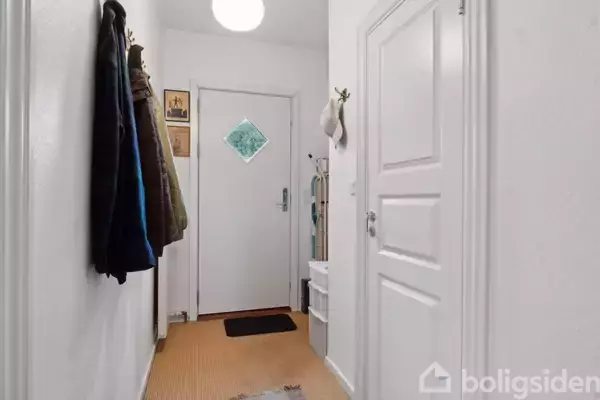 En gang med hængende jakker på venstre væg, en lukket dør foran og en strikket knagerække på højre væg. Lyse vægge og en lav, hvid skænk ved døren.