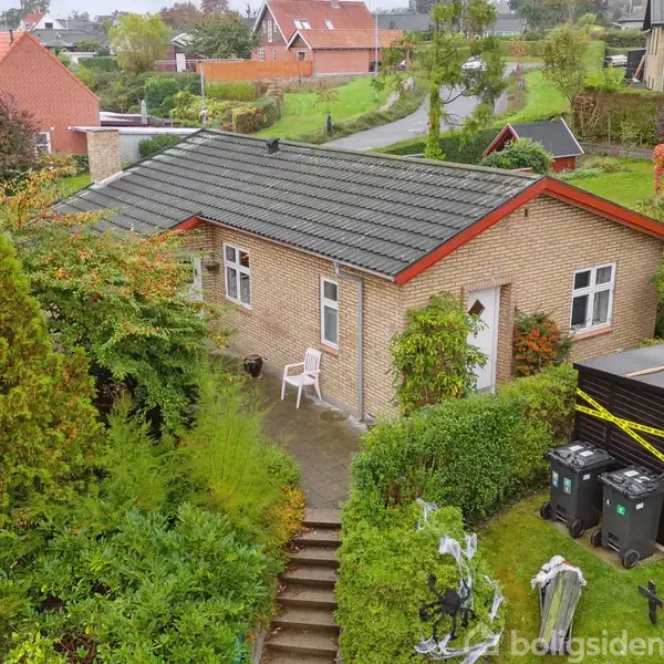 En etplans murstenshus med gråt tag står omgivet af frodig vegetation. Foran er en haveskur og affaldsbeholdere. Trappe fører op til indgangen. Omkringliggende kvarteret består af lignende huse.