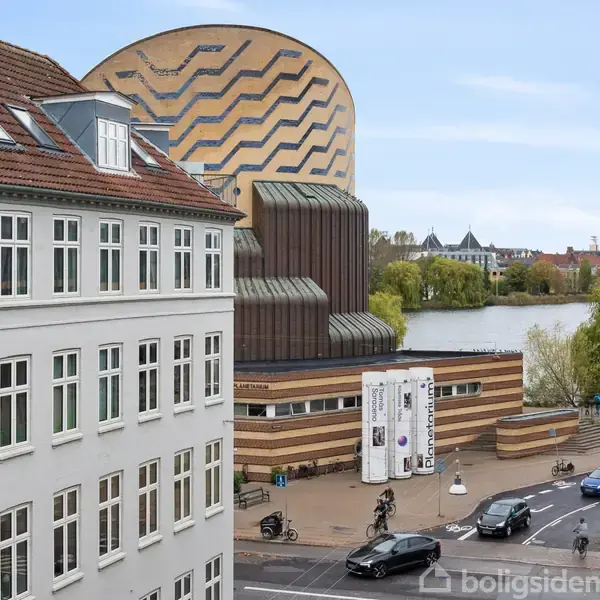 En hvid bygning står ved en vej hvor biler og cykler passerer. I baggrunden ses en moderne, bølget bygning nær en sø med træer og byudsigter.