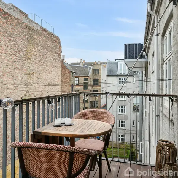 En altan med et rundt bord og to kurvestole placeret ved rækværket. Omgivet af murstensbygninger, ses små haver og himmel i baggrunden. En lyskæde hænger på rækværket.