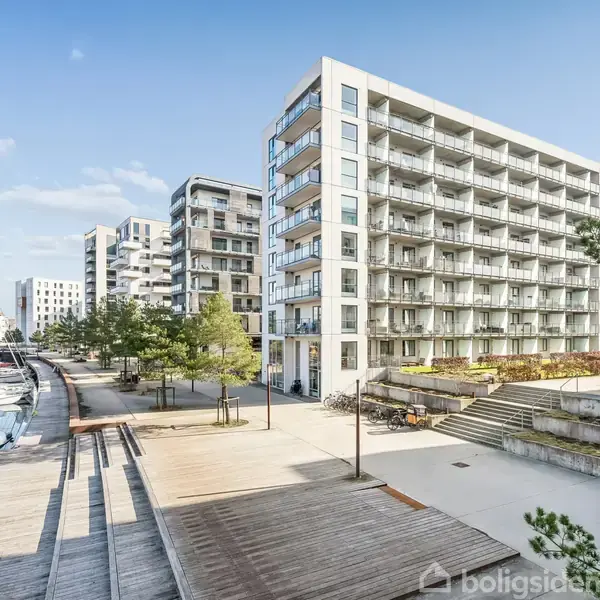 En moderne lejlighedsbygning står ved en havnepromenade med både, omkranset af træer og stier. Bygningen har flere altaner og er omgivet af en urban boligområde.