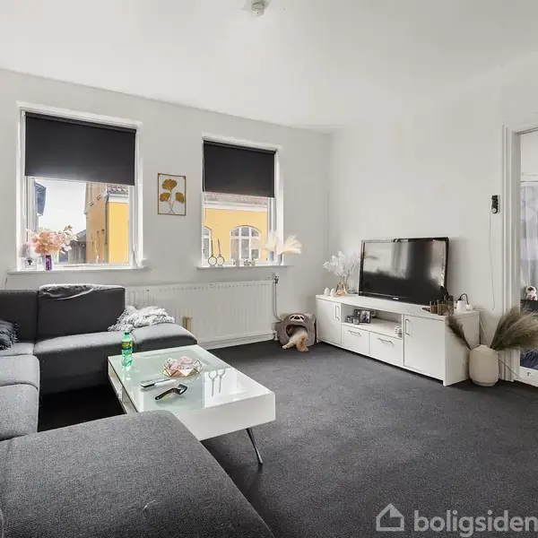 Gråt sofahjørne med sofabord, står i en lys stue med tv-bord og fladskærm. Vinduet har sorte rullegardiner, og der er døråbning til tilstødende rum.