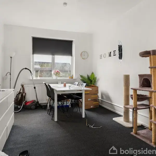 Et rum med et bord og stole i midten, omgivet af en høj katteklatretræ til højre. Væggen har dekorationer og ordet "HOME". Skuffer står ved siden af bordet.