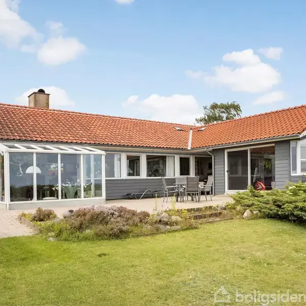 En moderne villa med rødt tegltag har en stor terrasse med spisebord og stole. Huset har en vinterhave med store vinduer. Det ligger i en frodig have med græsplæne.
