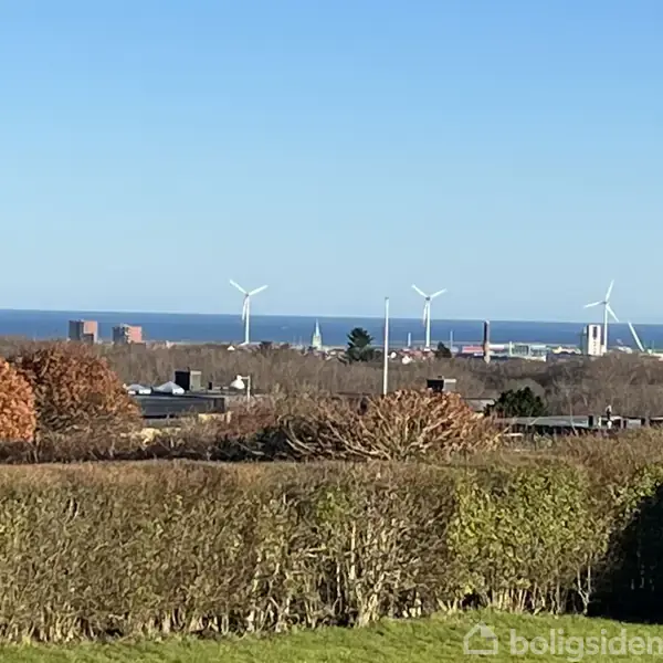 Vindmøller står stille i baggrunden, godt over en by med varieret arkitektur og farver, omgivet af frodige træer og buske under en skyfri blå himmel.