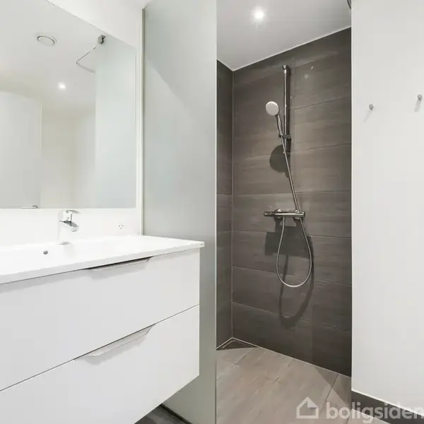 Håndvask med indbygget skab står i et lyst badeværelse. Ved siden af er en bruser monteret på en mørk flisevæg. Rummet er moderne og minimalistisk indrettet.