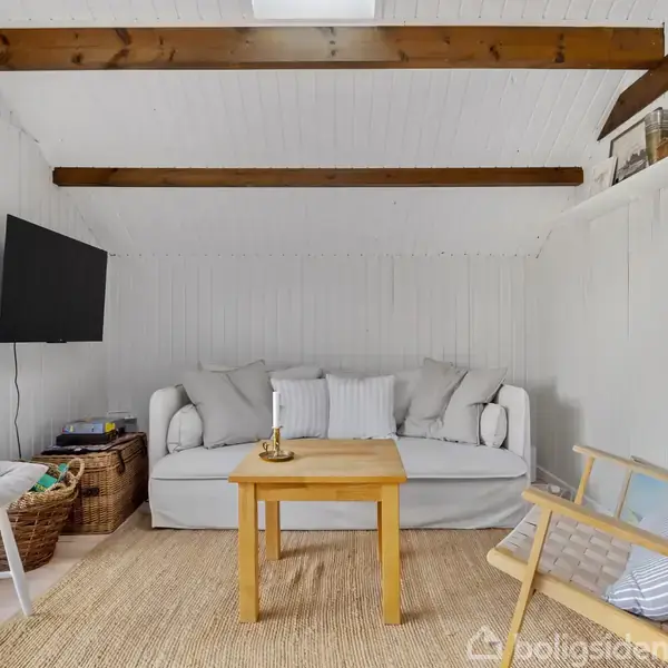 Sofa står i en lys stue med bjælkelofter. Foran sofaen er et træbord, omgivet af stole, en kurv og en TV-skærm på væggen. Hyggelig atmosfære.
