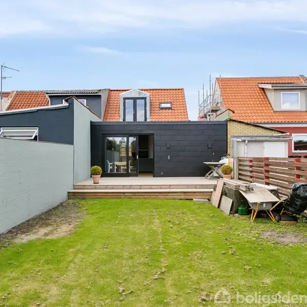 Hus med moderne tilbygning i sort facade med vinduer, står mellem to huse med røde tage. Foran er en grøn græsplæne omgivet af hegn og et lille trædæk.