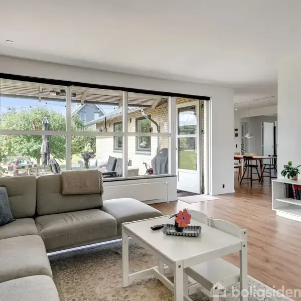 En stue med en grå sofa står stille ved et sofabord. Contextet består af en stor havedør, der åbner ud til en terrasse og have. Tv'et hænger på væggen.
